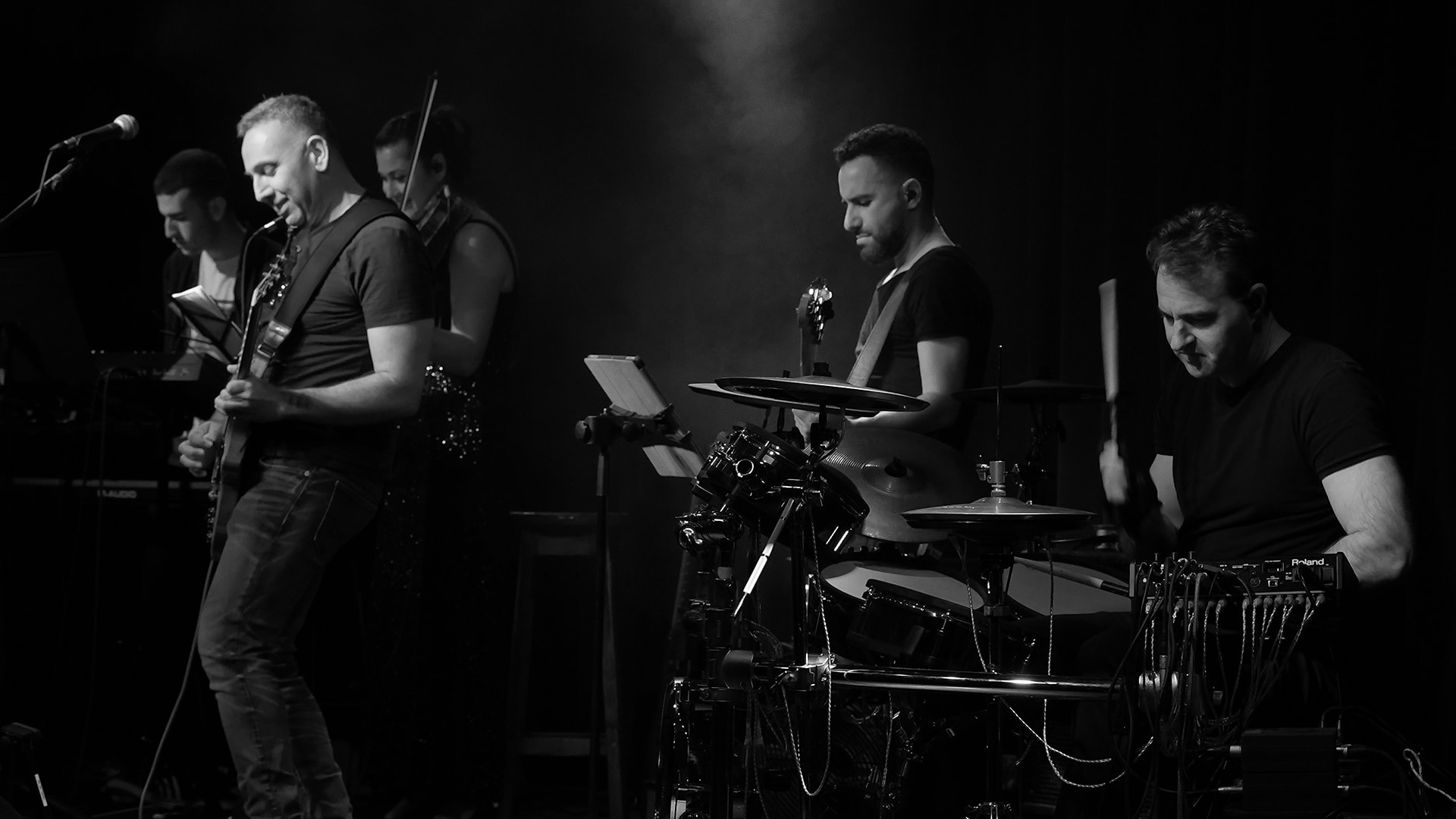 להקת הרוק קאדנס במהלך הופעה על הבמה בתל אביב. אמיר בתופים, רן גיטרה ושירה. יש גם כינור. בפילטר שחור.